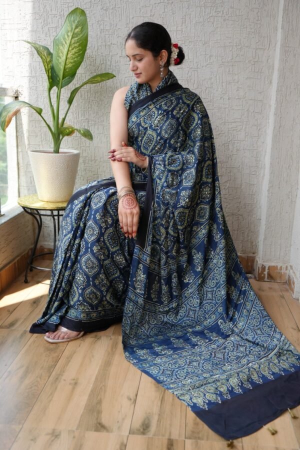 Woman draped in indigo blue Ajrakh print Modal silk saree with navy border, seated near a plant